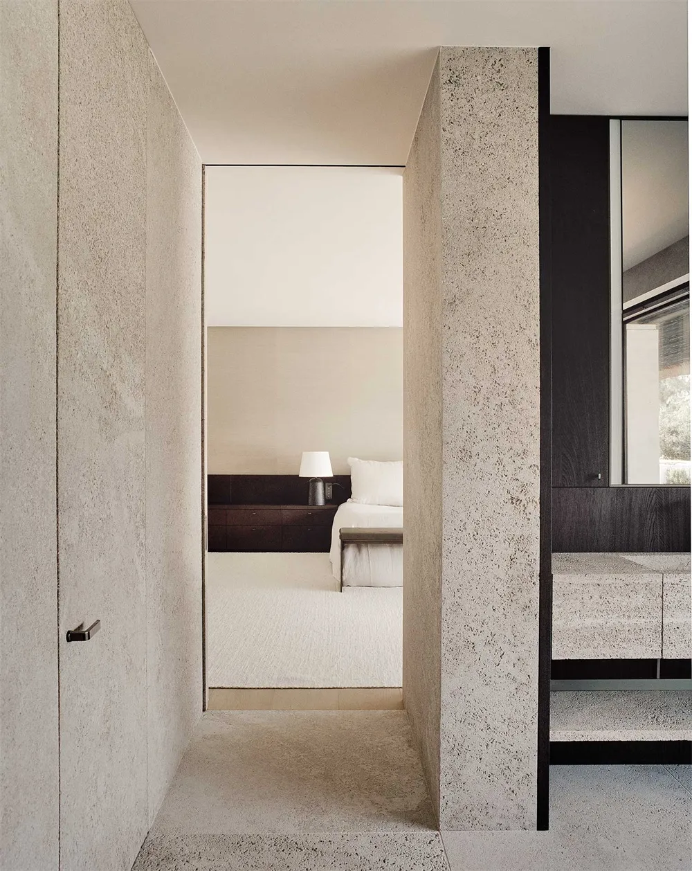 Minimalist bedroom with concrete walls and floor, featuring a bed with white sheets and pillows, and a nightstand with a lamp