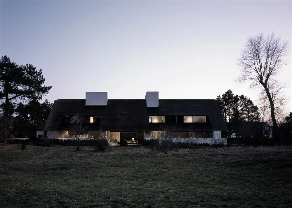A modern rural house with a thatched roof at night, featuring illuminated windows and a minimalist design.