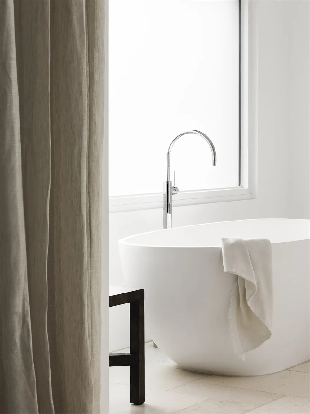 A minimalist bathroom featuring a ceramic freestanding bathtub, a towel draped over the side, a wooden stool, and a large win