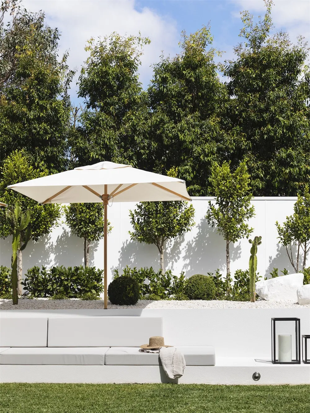 A minimalist outdoor patio with a white concrete seating area, a large white umbrella, and greenery in the background.
