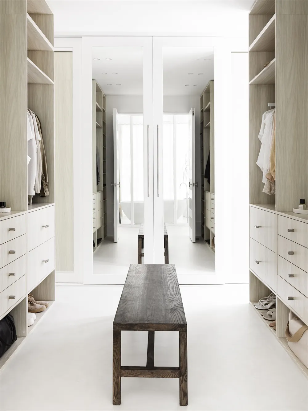 A modern white closet with open shelves, a wooden bench, and two large mirrors.