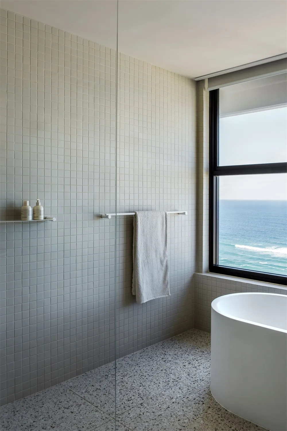 Modern bathroom with white tile walls and floor, featuring a window overlooking the ocean, a freestanding bathtub, and minima