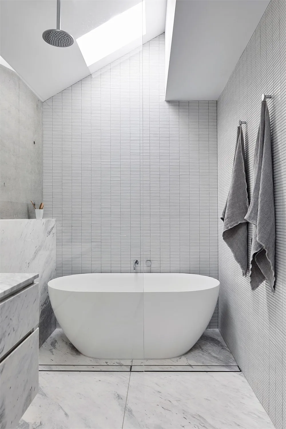 A minimalist bathroom with marble tiles, concrete walls, and a freestanding bathtub under a skylight.