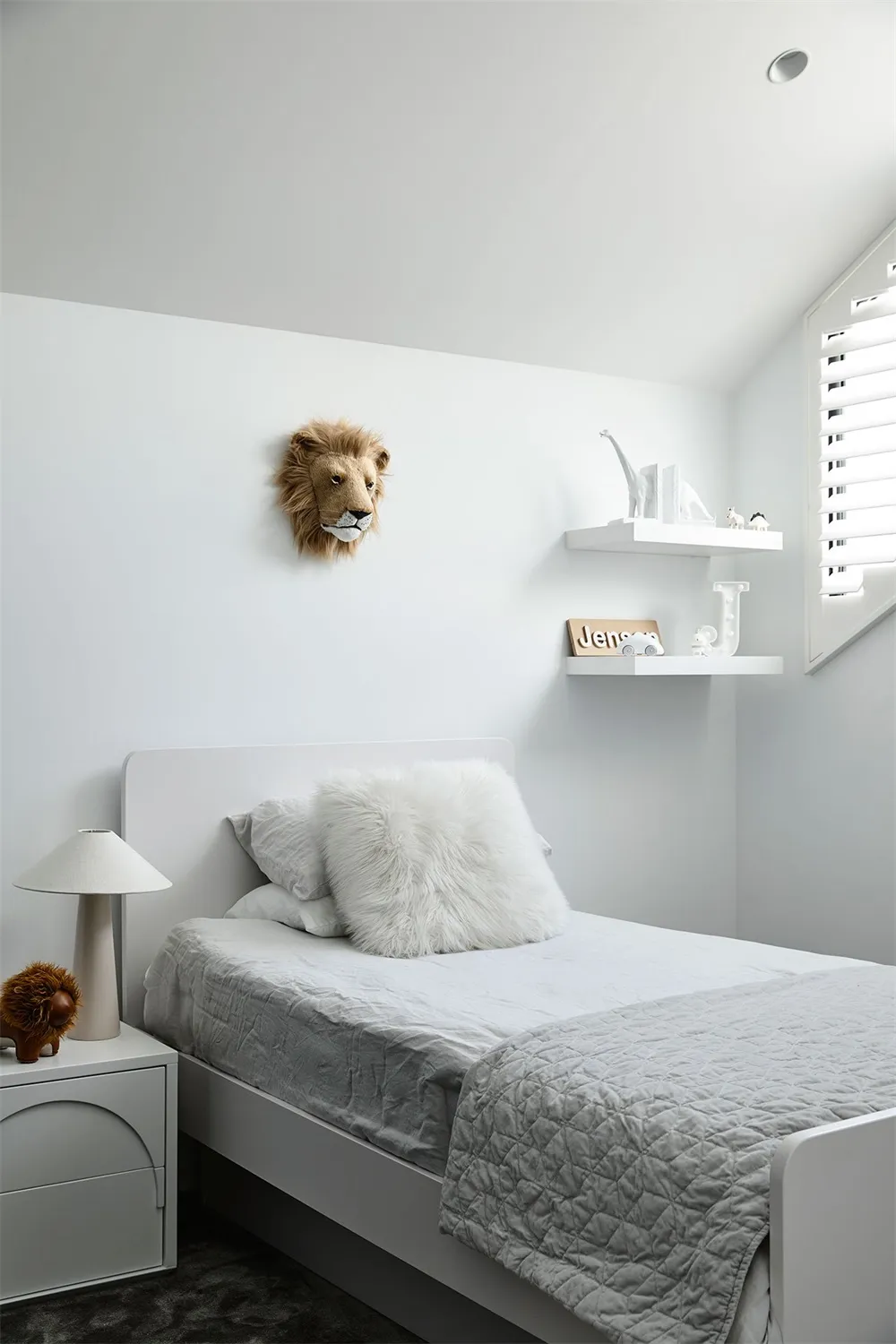A minimalist bedroom with a white bed, fur pillow, and wooden headboard, featuring a lion's head and a shelf with animal figu