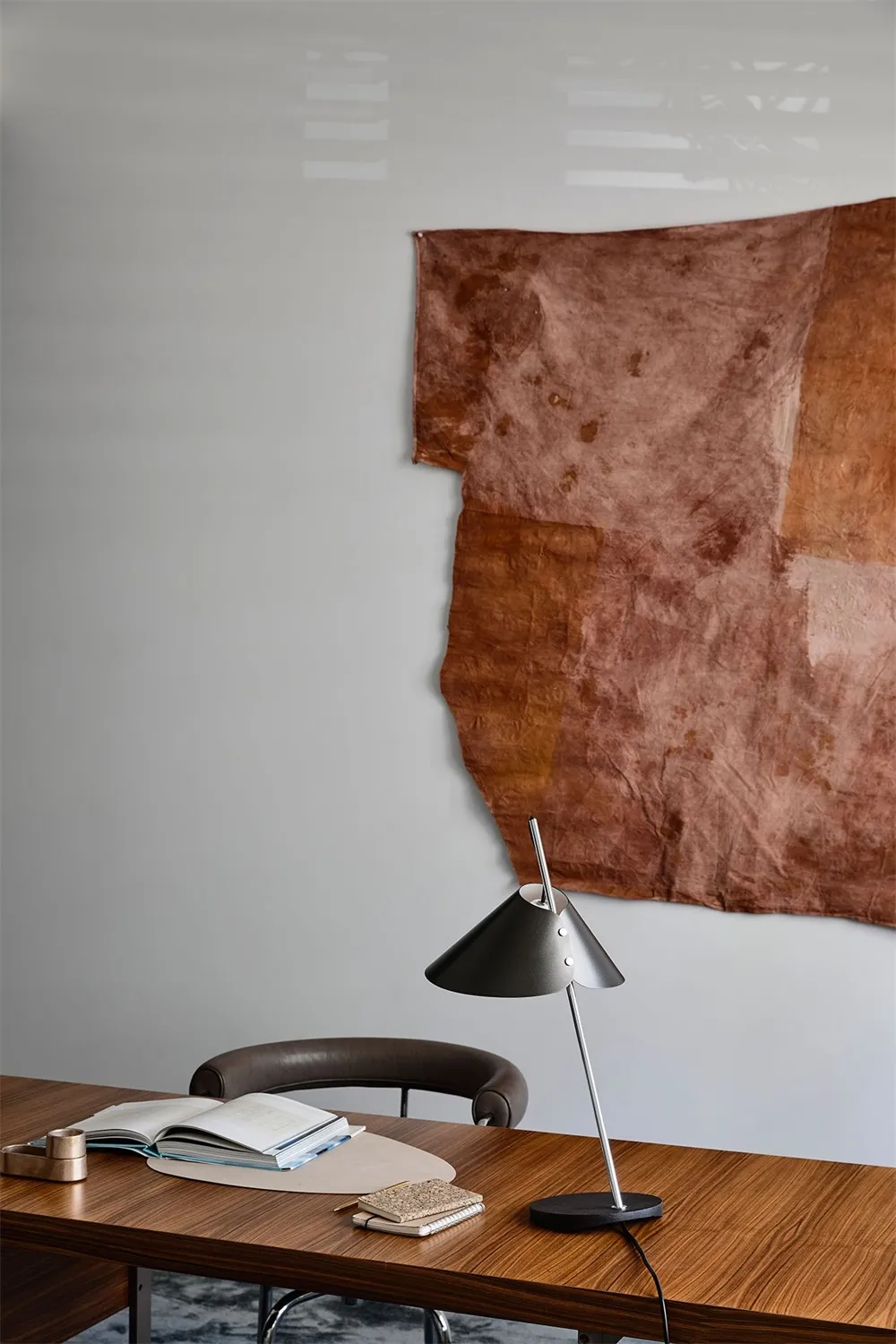 A minimalist workspace featuring a wooden desk, chair, and lamp, with a brown fabric hanging on the wall.