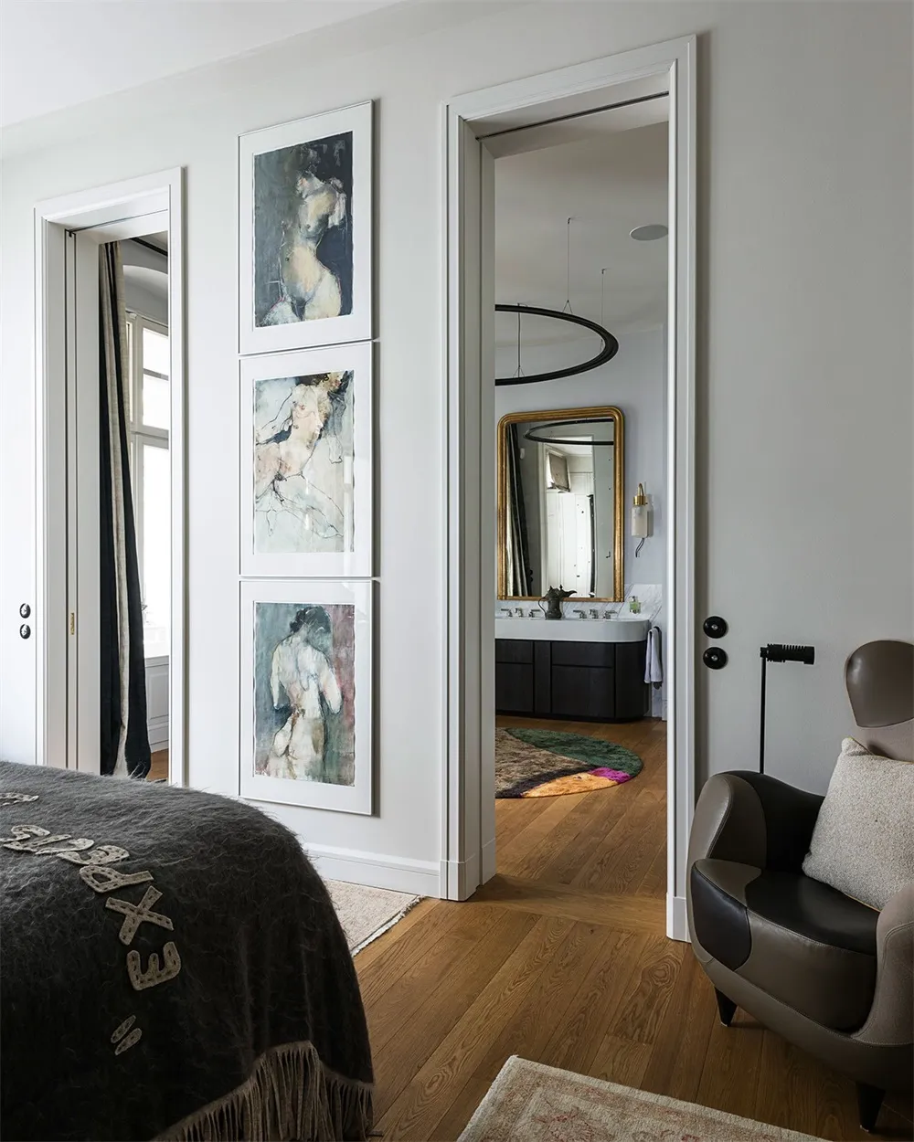 A modern bedroom featuring wooden flooring and a carpet, with a door leading to a bathroom and an armchair in the foreground.