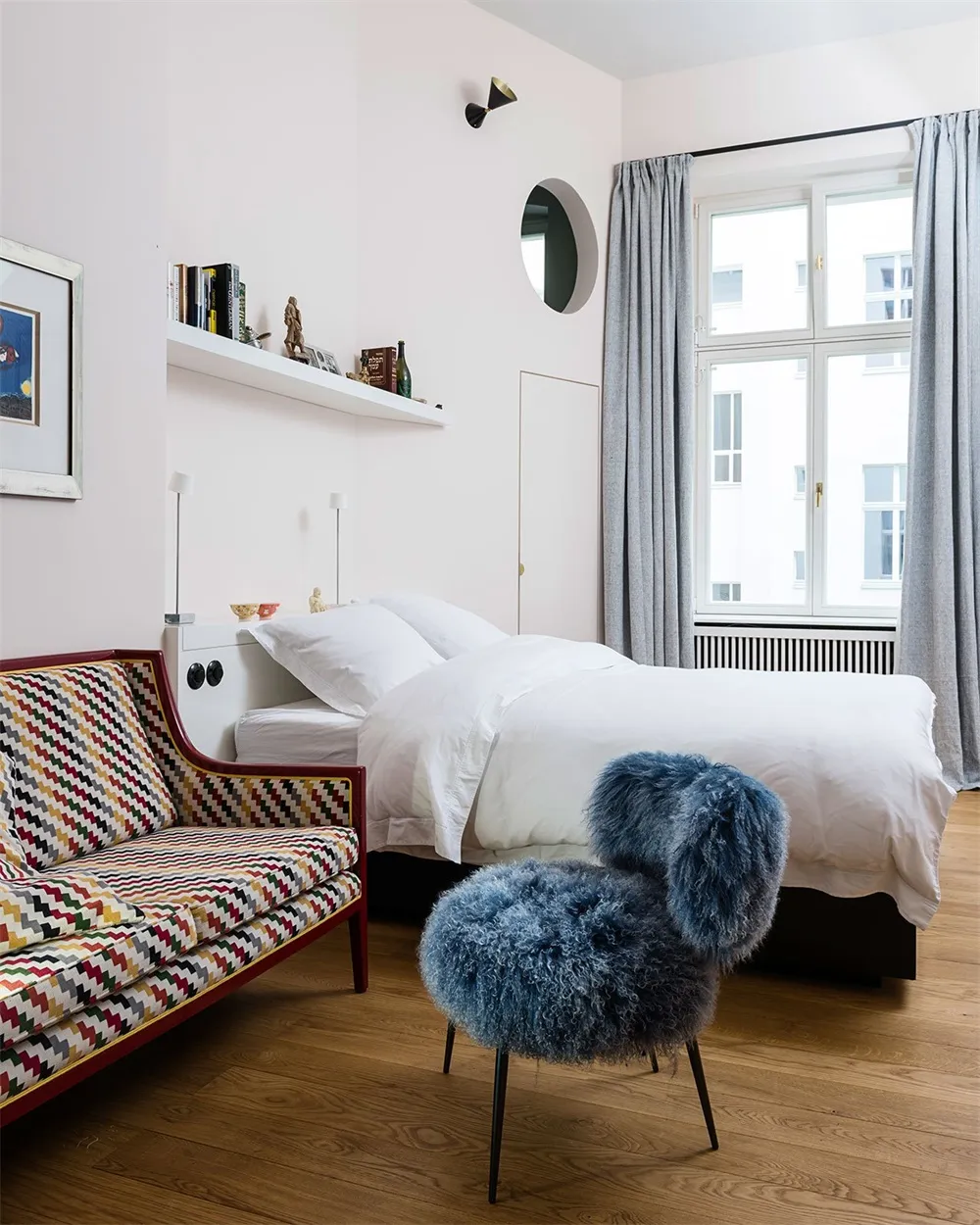 A modern bedroom with a white bed on a wooden floor, featuring a colorful couch and fluffy chair, and a shelf with decorative