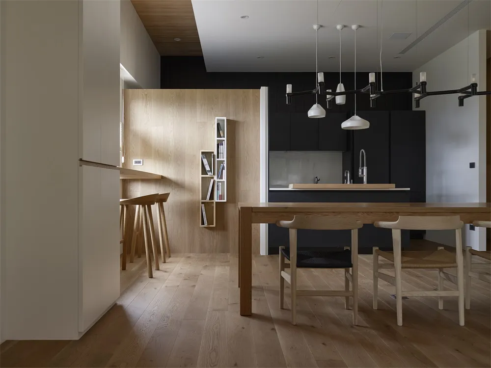 Modern, minimalist kitchen with wooden flooring, cabinetry, and furniture. Features sleek, black countertops and pendant ligh