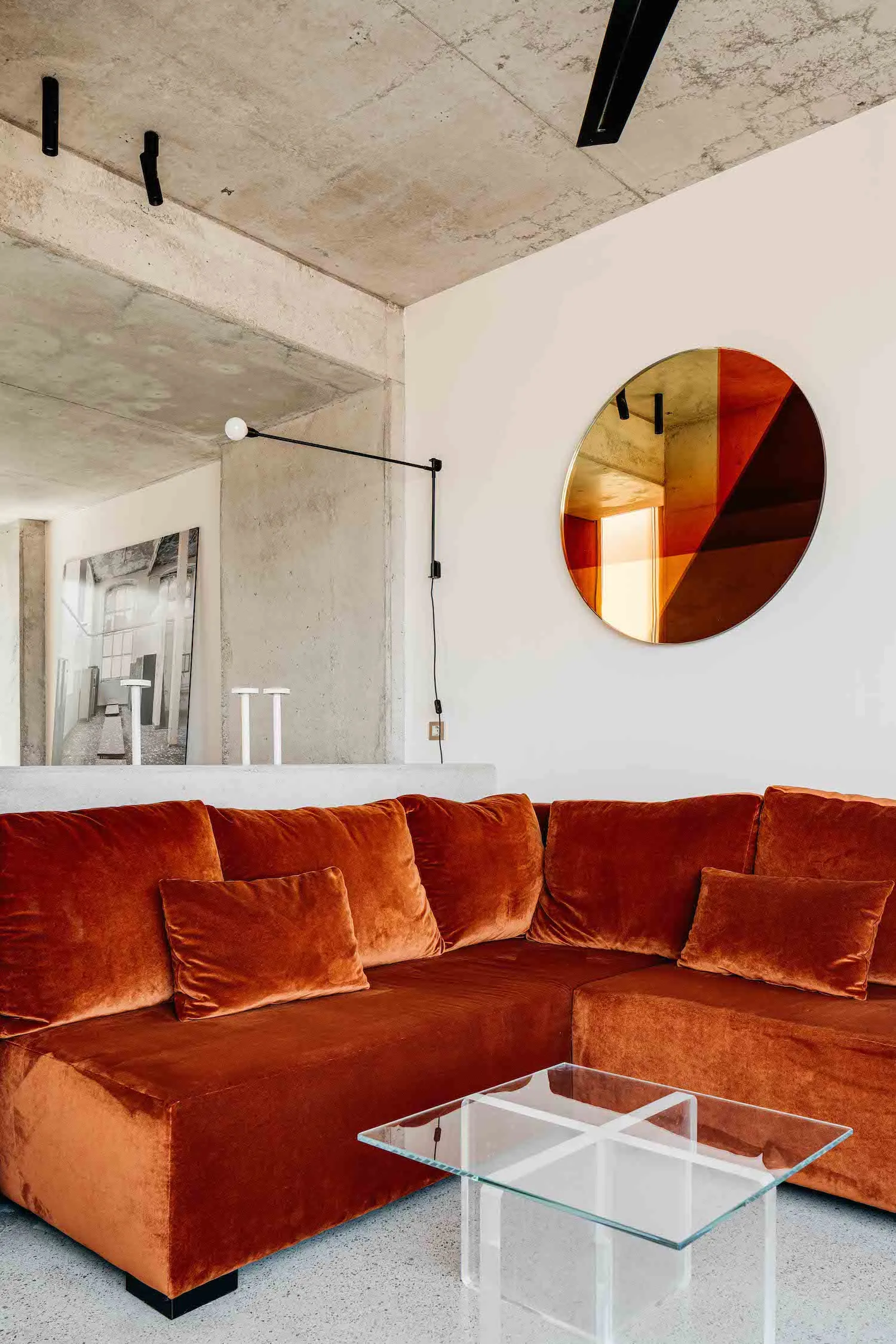 Modern living room with an orange velvet sofa, concrete walls, and a circular metallic mirror.