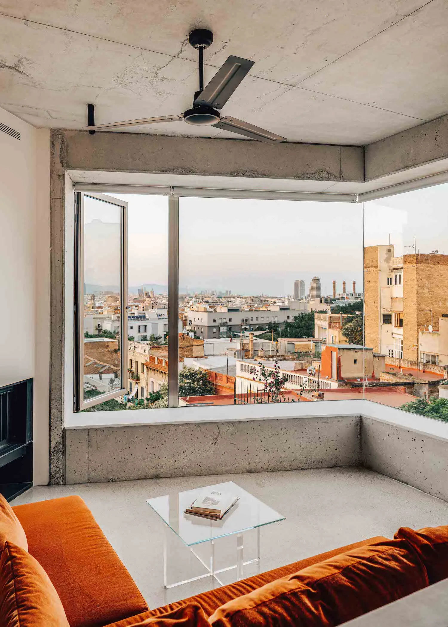 Modern minimalist living room with concrete walls, glass table, orange couch, and cityscape view through large windows.