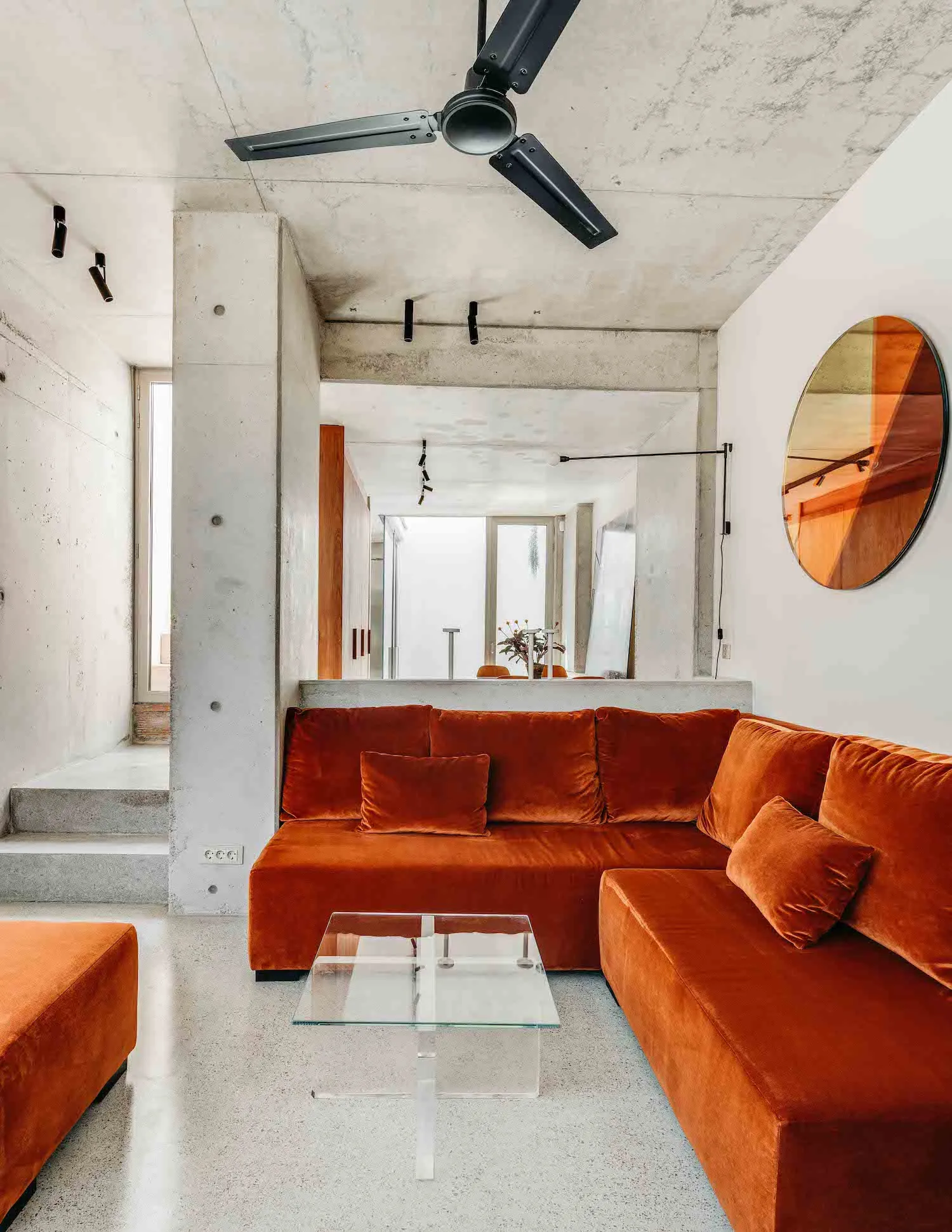 A modern industrial living room with concrete walls, ceiling fan, and a large terracotta sofa.