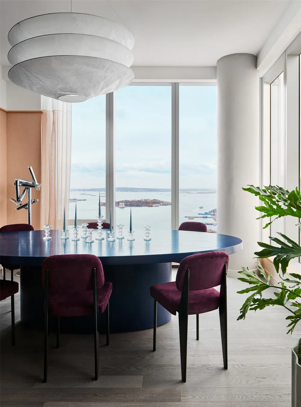 A modern dining room features a round wooden table with purple upholstered chairs, glass candle holders, and large windows ov