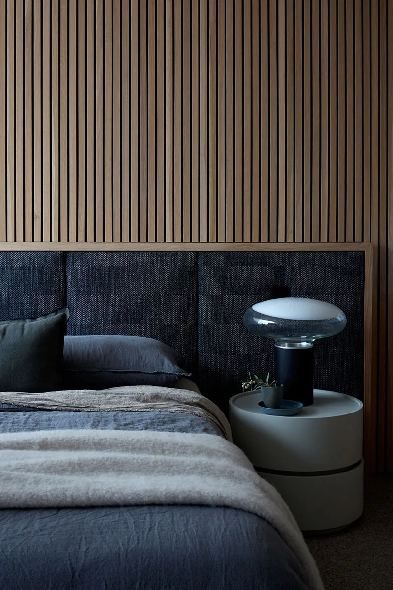 A modern bedroom featuring a bed with a wood-paneled headboard, grey linens, and a bedside table with a lamp and plant.