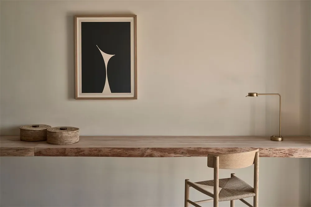 A minimalist home office with a wooden desk, chair, and lamp, featuring a black and white abstract artwork on the wall.