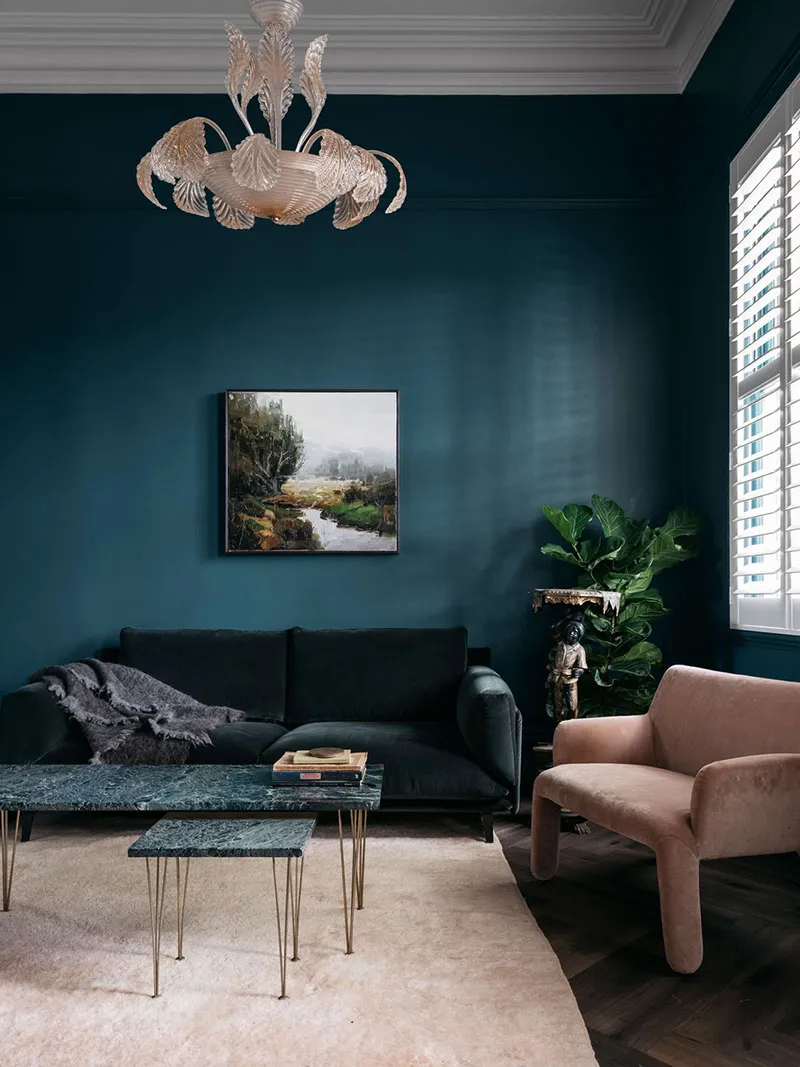 A modern living room featuring a dark green velvet sofa, a marble coffee table, and a pink armchair. The room has dark teal w