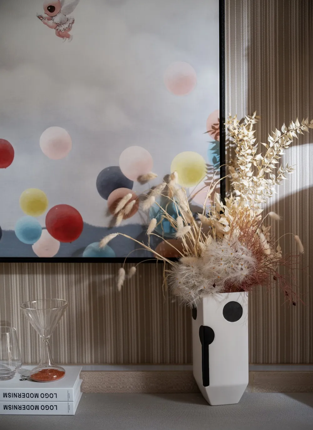 A decorative arrangement featuring a white vase with black dots and various dried flora against a striped background with a c