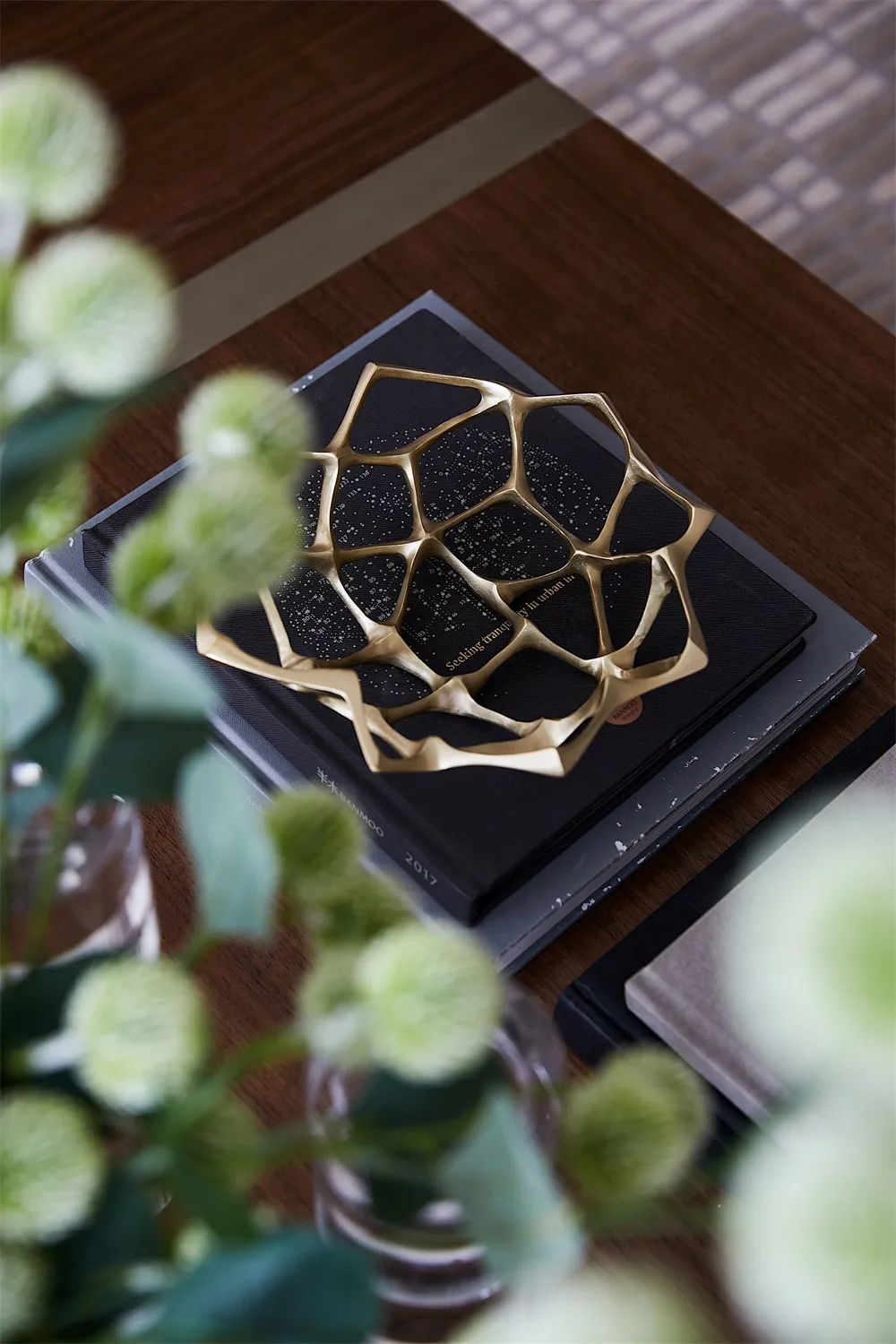 A metallic, geometric, modern decorative object with a star-like shape placed on top of a stack of black books on a wooden su
