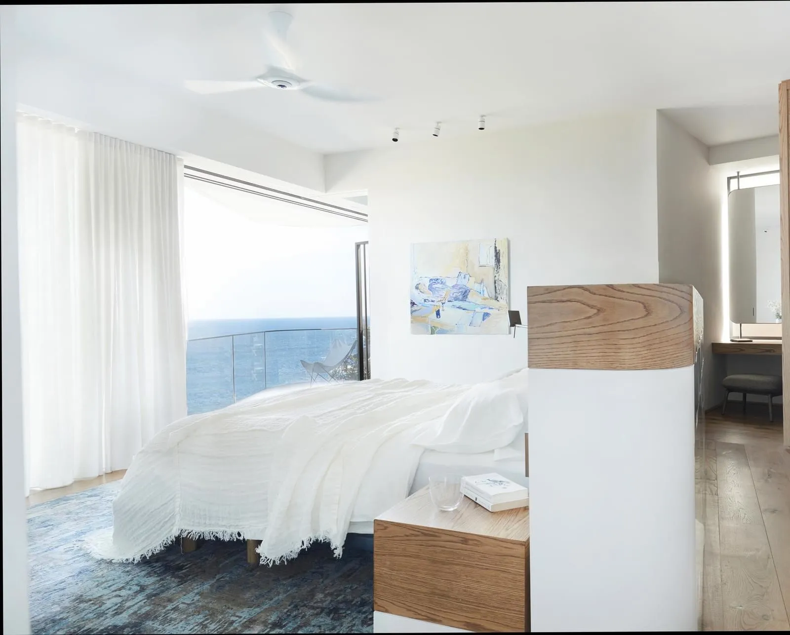 A modern minimalist bedroom featuring white bedding, a wooden nightstand, and a white accent wall with a small abstract paint