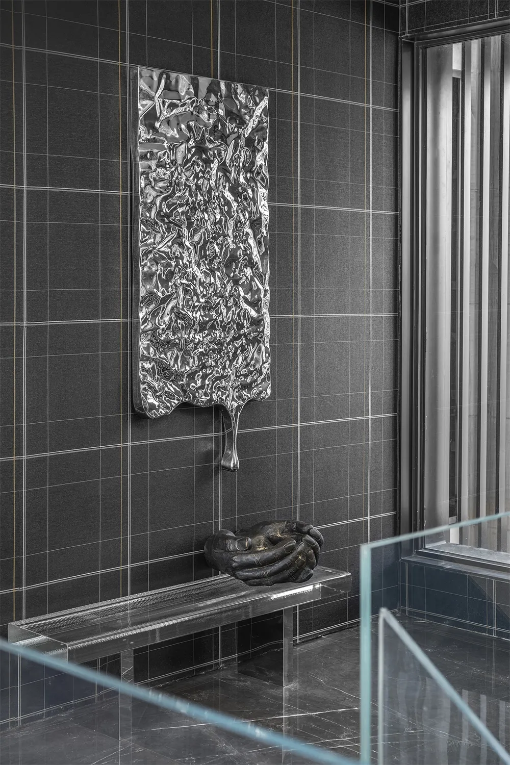 A metallic panel with a crumpled surface texture is mounted on a dark grid-tiled wall, accompanied by a sculptural hand gestu