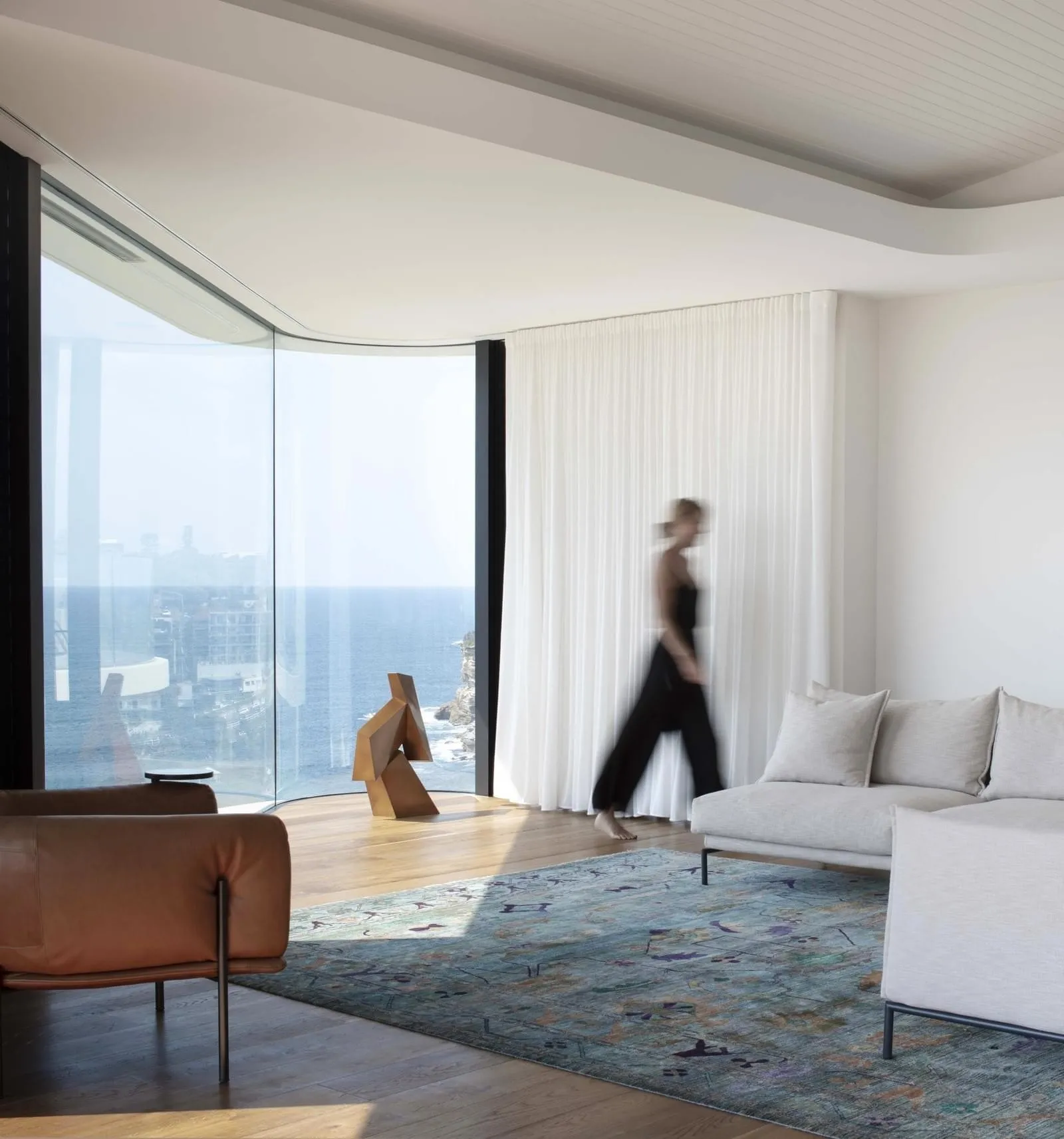 A modern minimalist living room with a sea view, featuring a white sofa, a brown leather armchair, and a patterned rug.