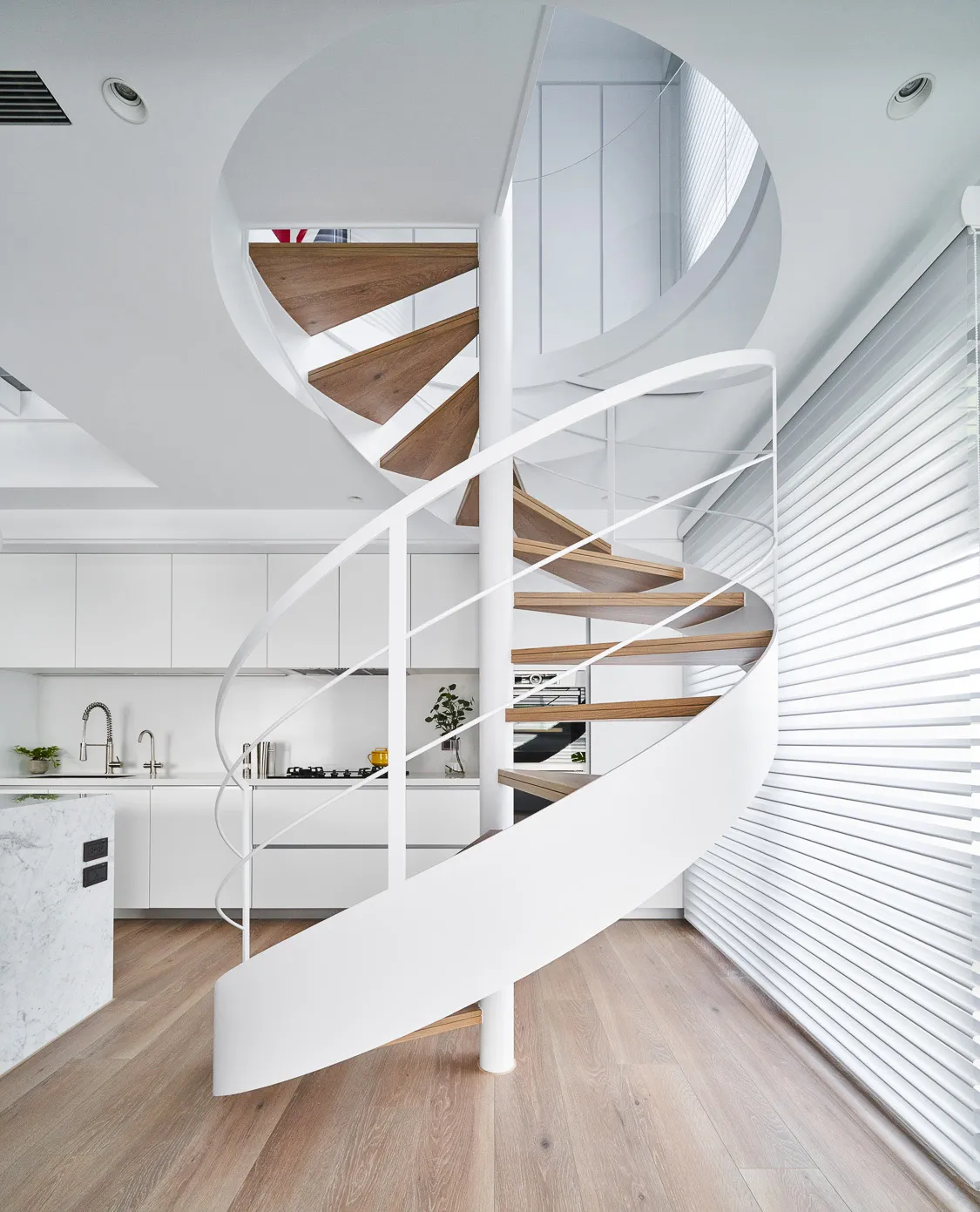 A modern, minimalist kitchen featuring a spiral staircase with white metal railings and wooden steps, designed with sleek, fu