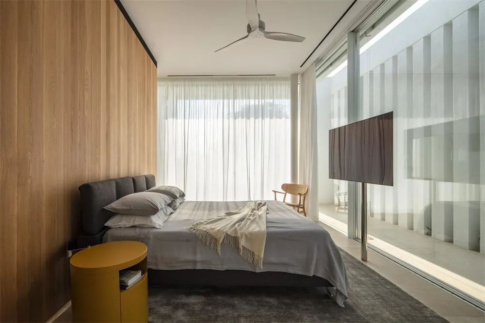 A modern minimalist bedroom featuring a wooden wall and floor, large glass windows, and a comfortable bed with grey bedding a