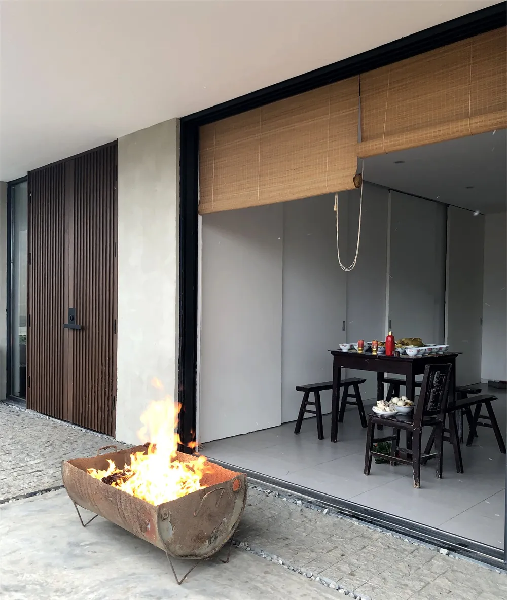 An industrial-style fire pit outdoors with flames burning, featuring a rustic metal container and a residential exterior with