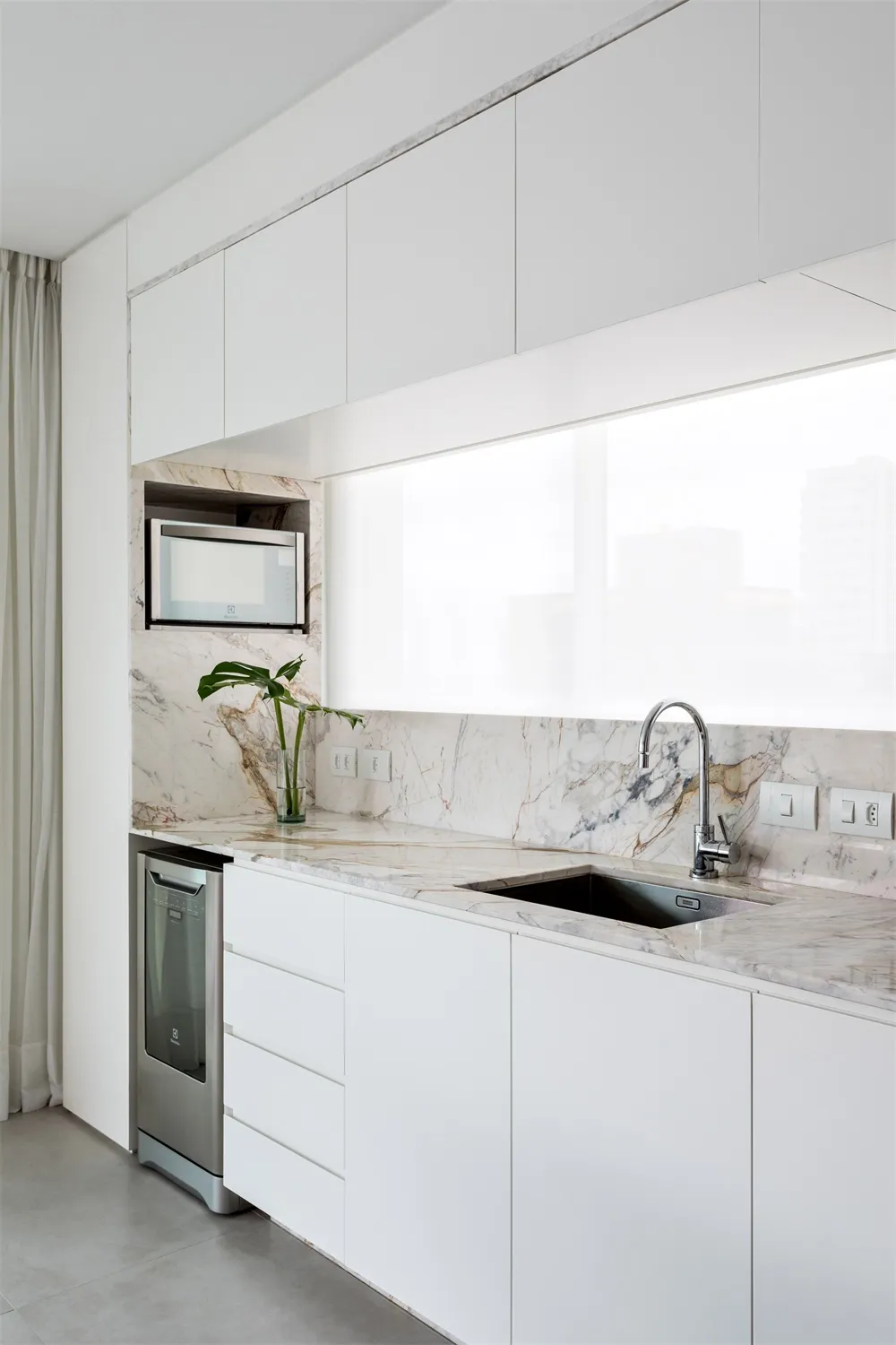 A modern, industrial residential kitchen with marble countertops and white cabinetry, featuring a sink, microwave, and minima