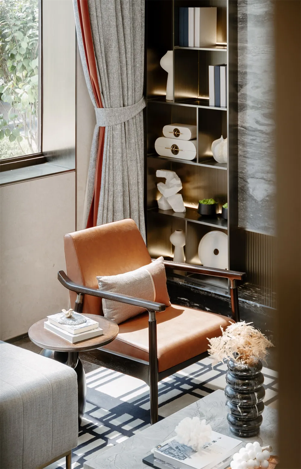 A modern living room featuring a leather armchair, a side table with stacked books, and a bookshelf with decorative items.
