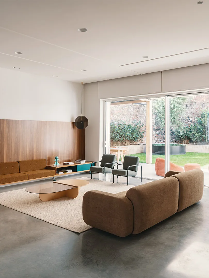 A modern living room with concrete flooring, a brown sectional sofa, a round glass coffee table, and a wooden shelf with deco