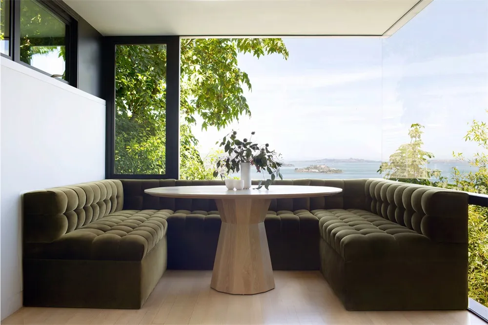 Modern living room featuring a velvet couch, round table, and large windows overlooking greenery.