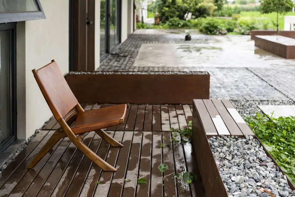 A modern wooden porch with a single chair and a bench, featuring a stone and gravel planter.