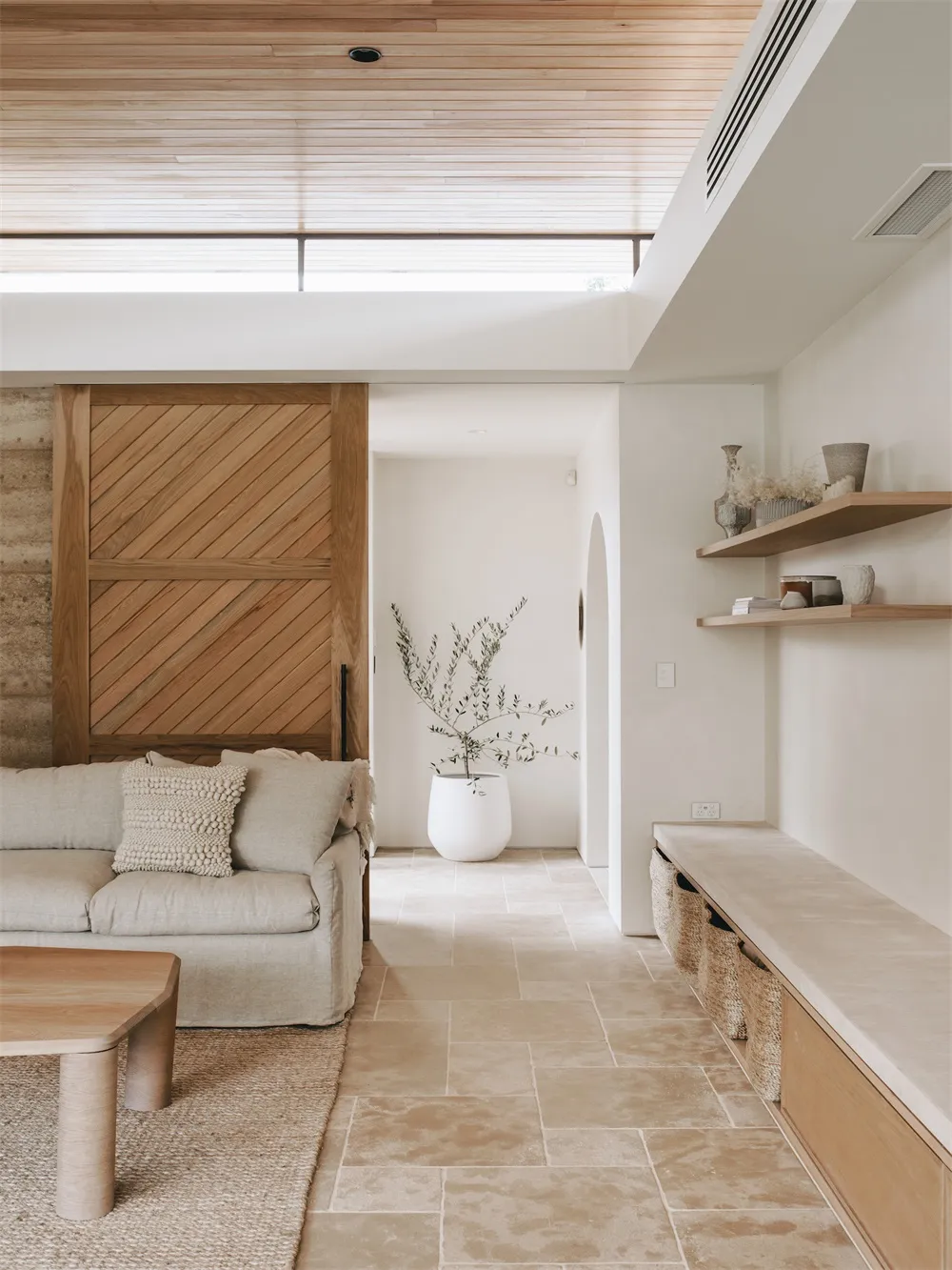 A minimalist living room featuring a beige couch, a wooden table, and a concrete floor. The room has a sliding wooden door an