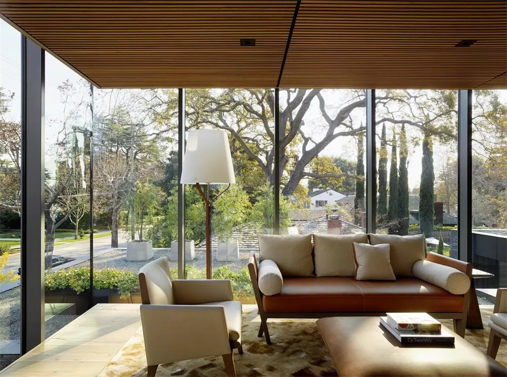 A modern living room featuring wooden walls and a brown leather sofa, with a large window showcasing a serene outdoor landsca