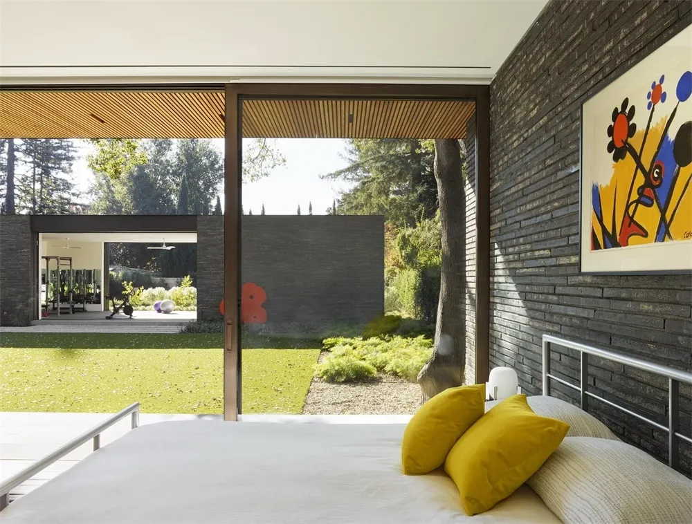 Modern bedroom with brick wall, yellow pillows, and white sheets, featuring a view of a garden with green grass and trees.