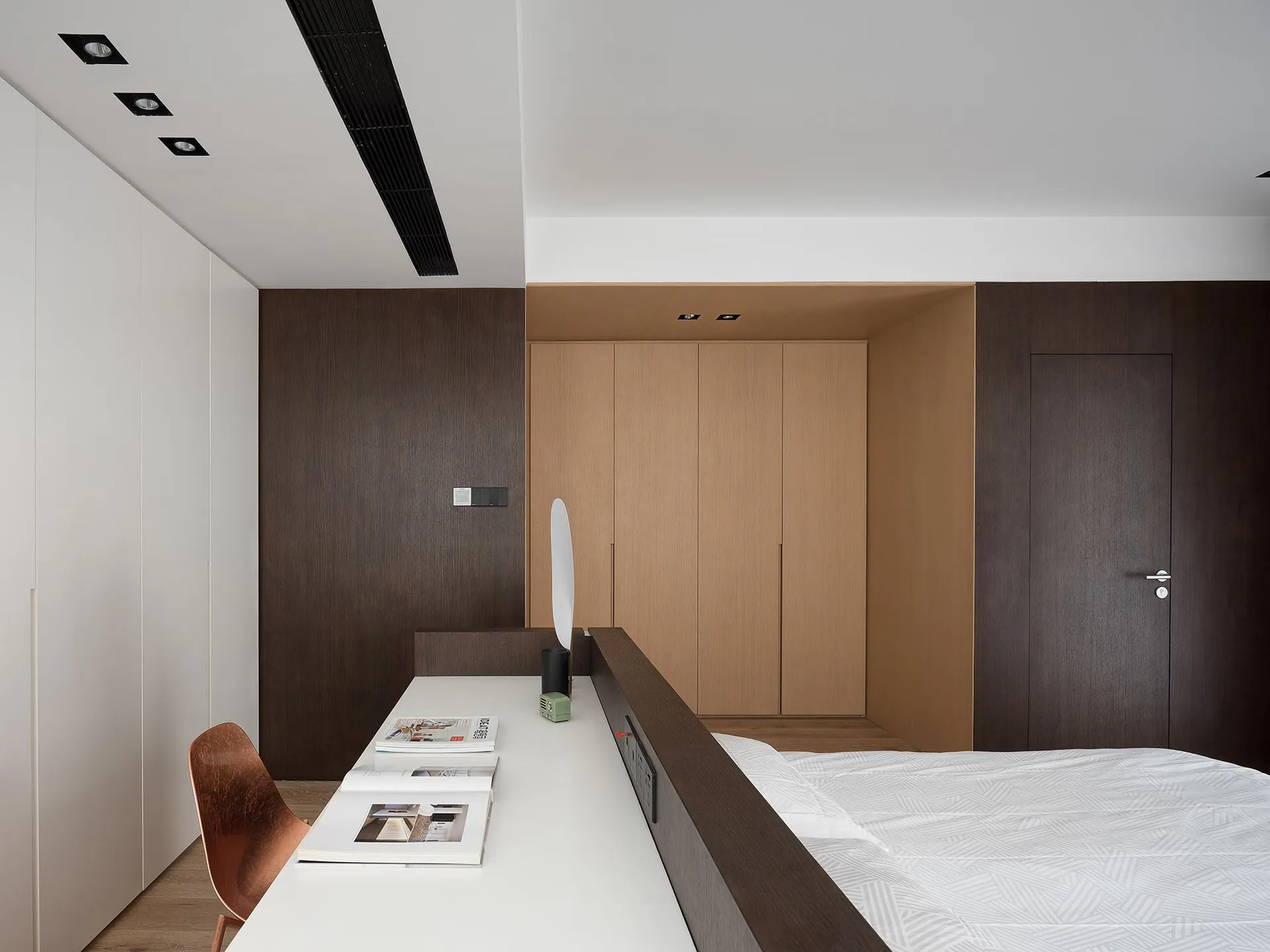 A minimalist bedroom with brown walls, a bed, and a white desk with a desk lamp.