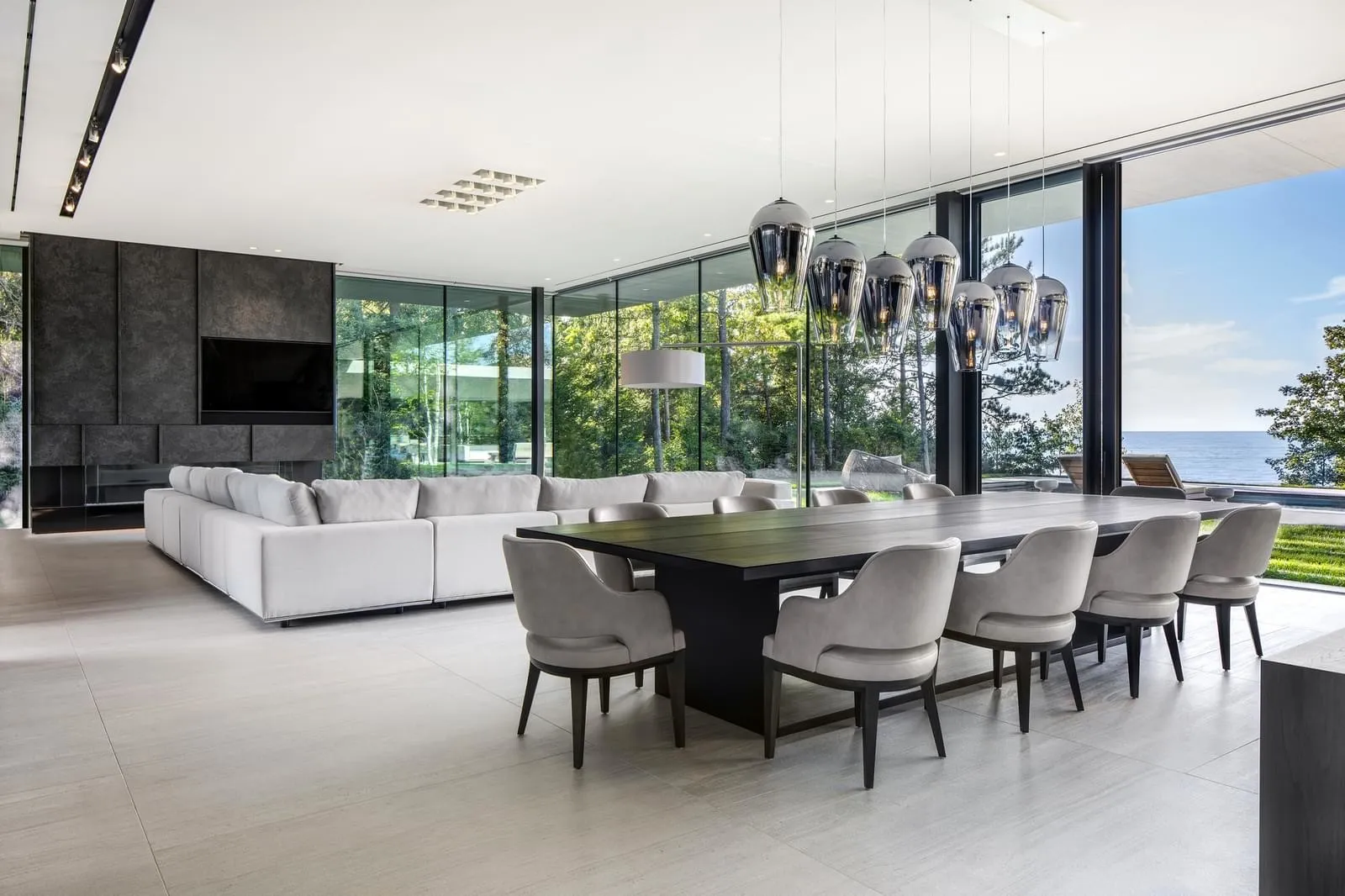 A modern living and dining room with white furniture, wooden table, and large windows overlooking the sea.
