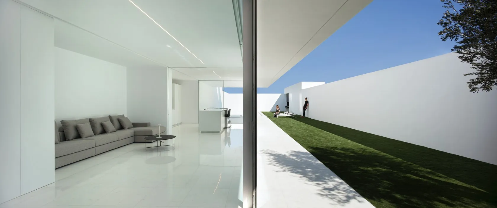 A minimalist residential exterior with glass walls, featuring a clean, modern design with white walls and a green lawn.