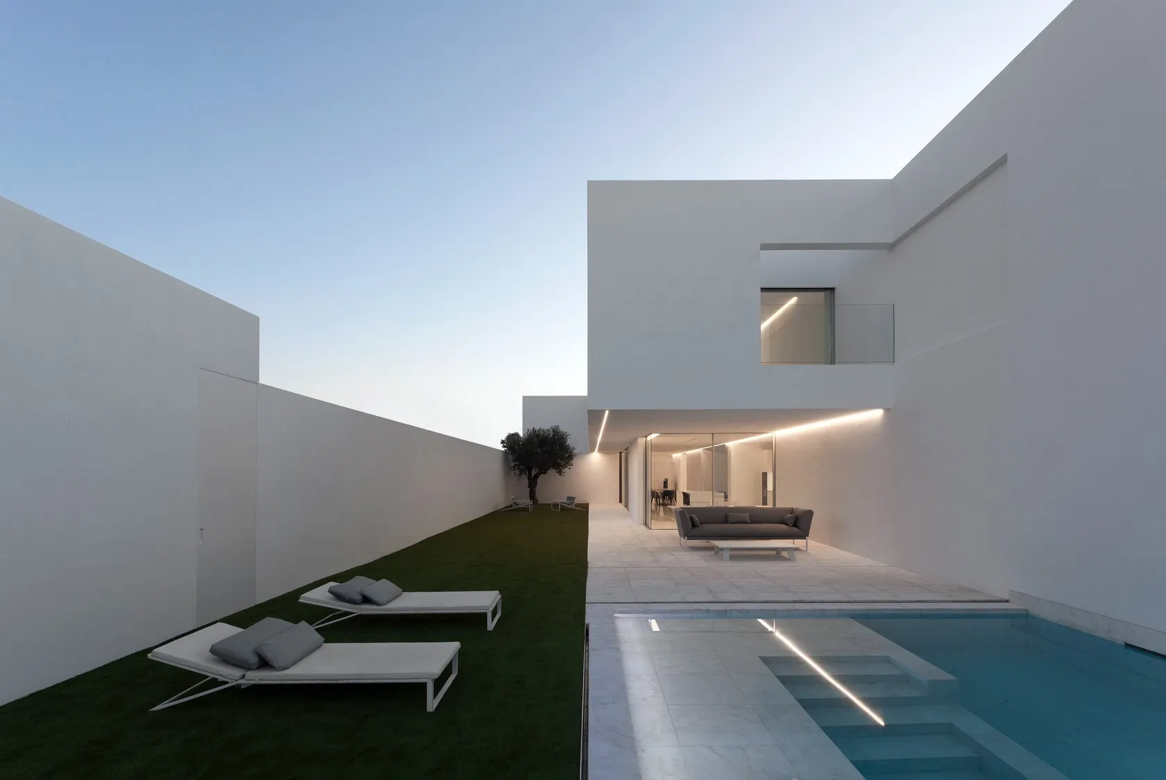A modern residential outdoor space with concrete elements, featuring a pool, lounge chairs, and a pathway leading to a house