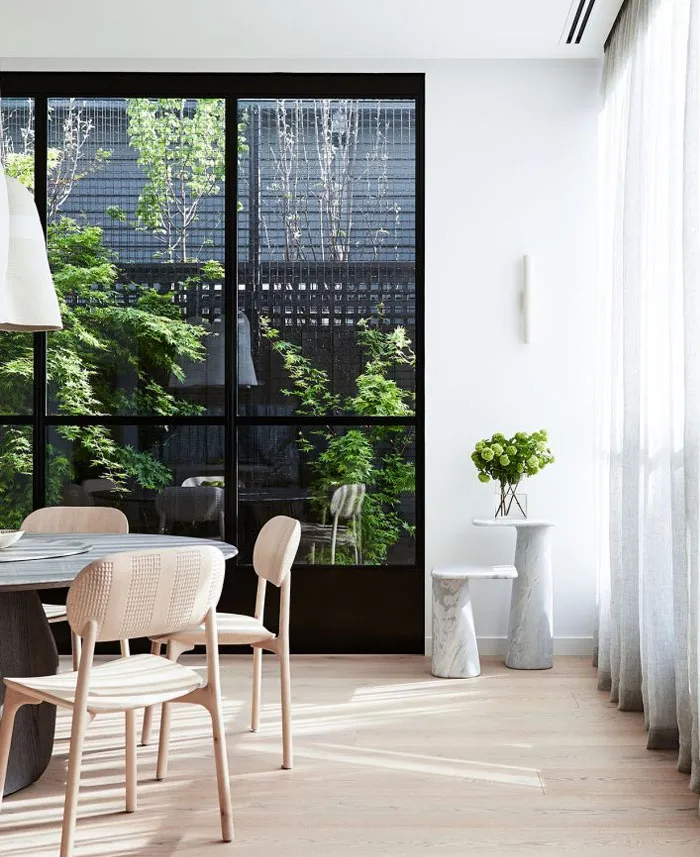 A minimalist dining room featuring wooden chairs and a marble table, surrounded by large windows.