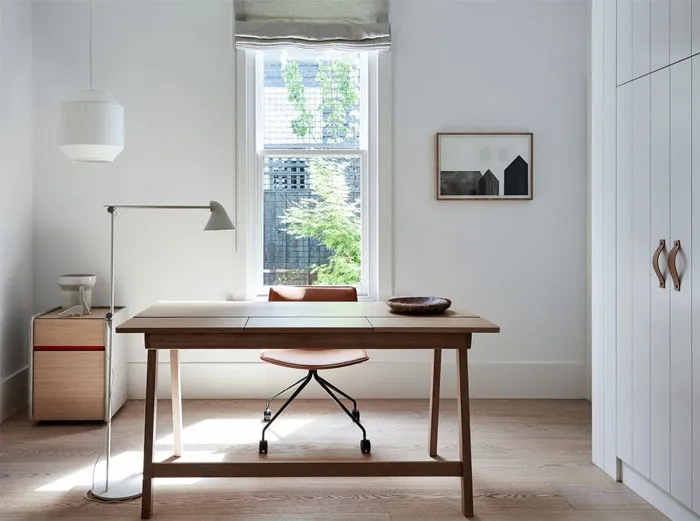 A minimalist home office featuring a wooden desk, modern chair, and metal cabinet seen through a window with greenery outside
