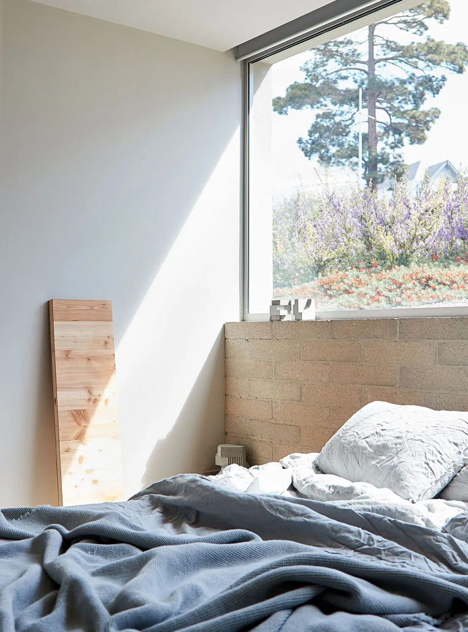 A modern bedroom with a concrete bed frame and large window overlooking a garden.