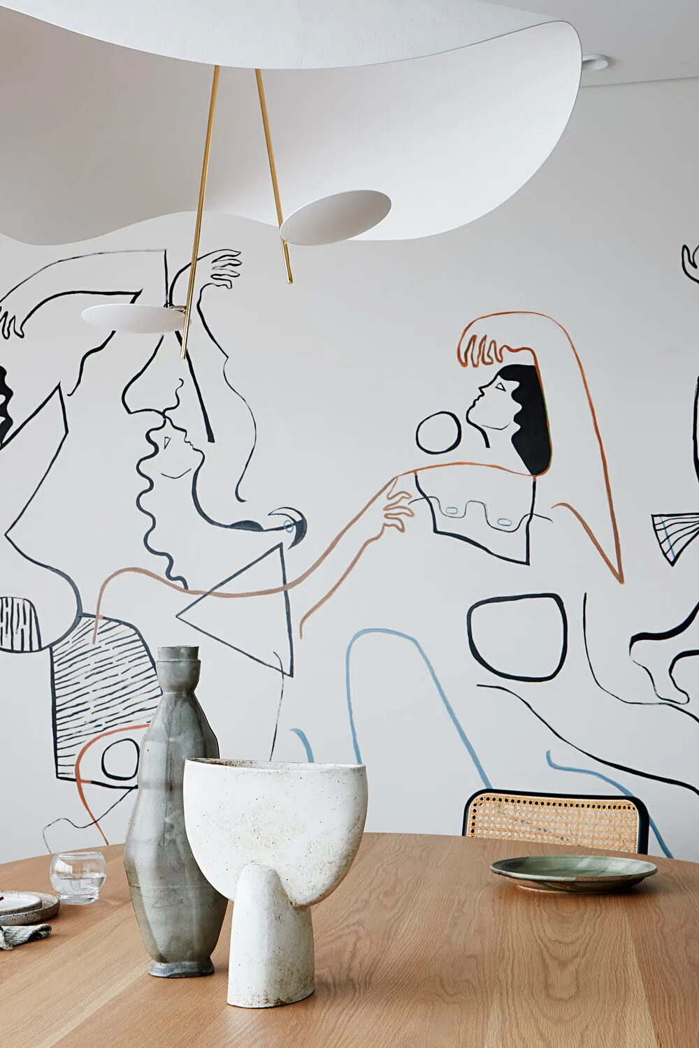 A modern dining room featuring ceramic tableware and a mural of abstract figures on the wall.