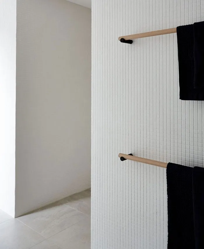 A modern minimalist bathroom with white ceramic tiles and black towels hanging on a wooden rack.