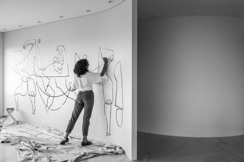 A woman artist painting minimalist figures on a white wall.