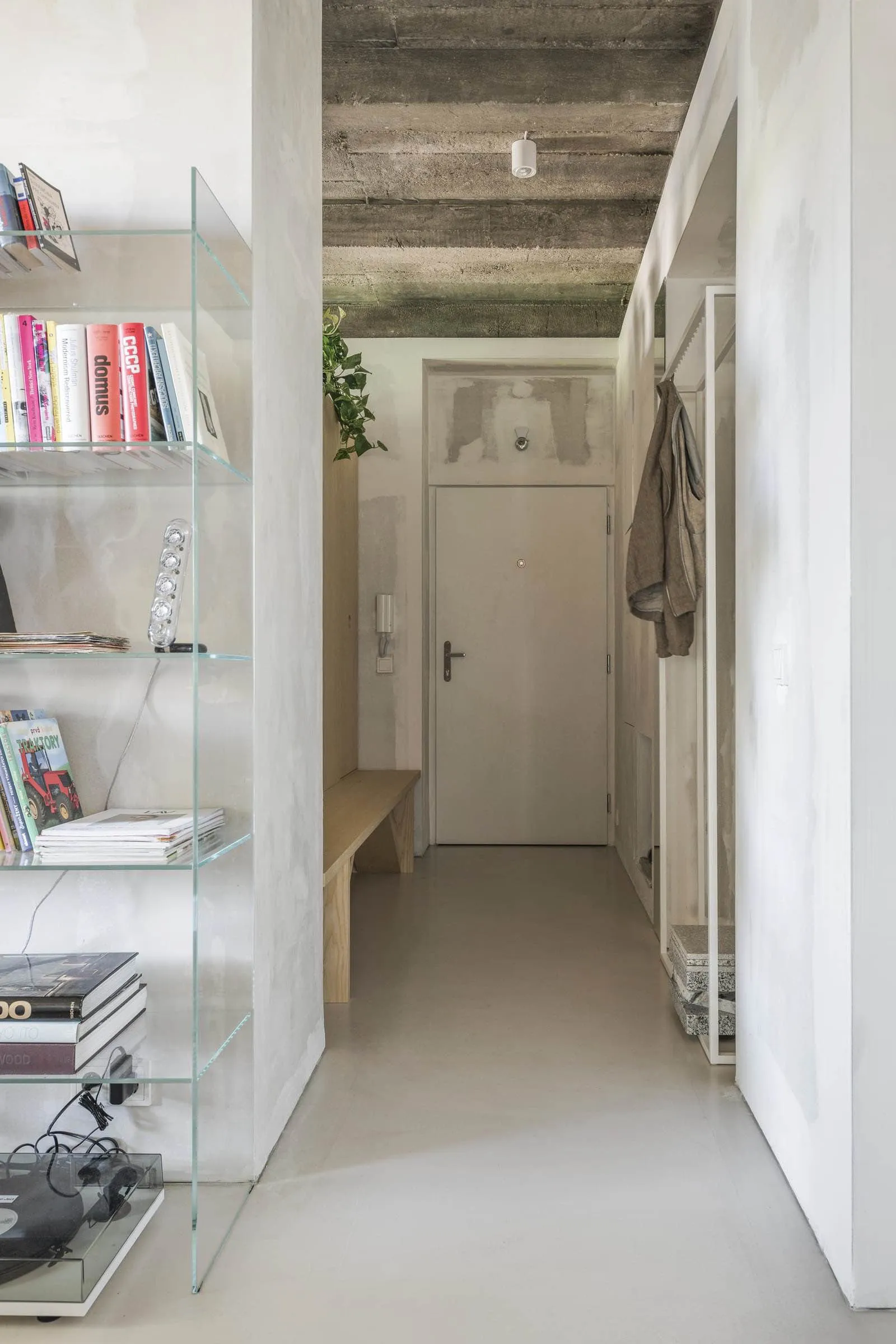 Modern minimalist entryway with wooden bench, glass bookshelf, and concrete ceiling.