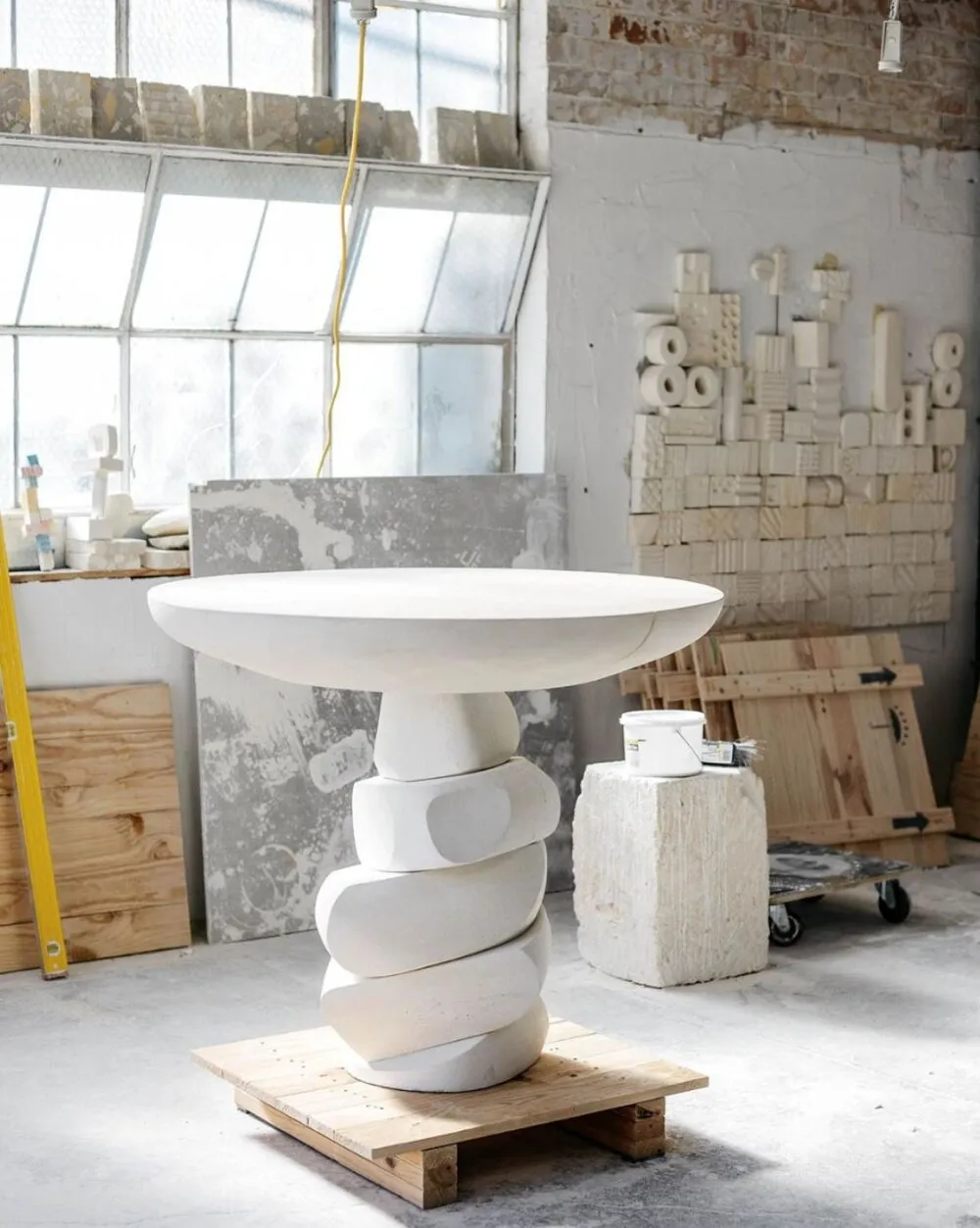 A modern, white plaster side table with a unique, stacked design placed on a wooden pallet in an indoor workspace with scatte