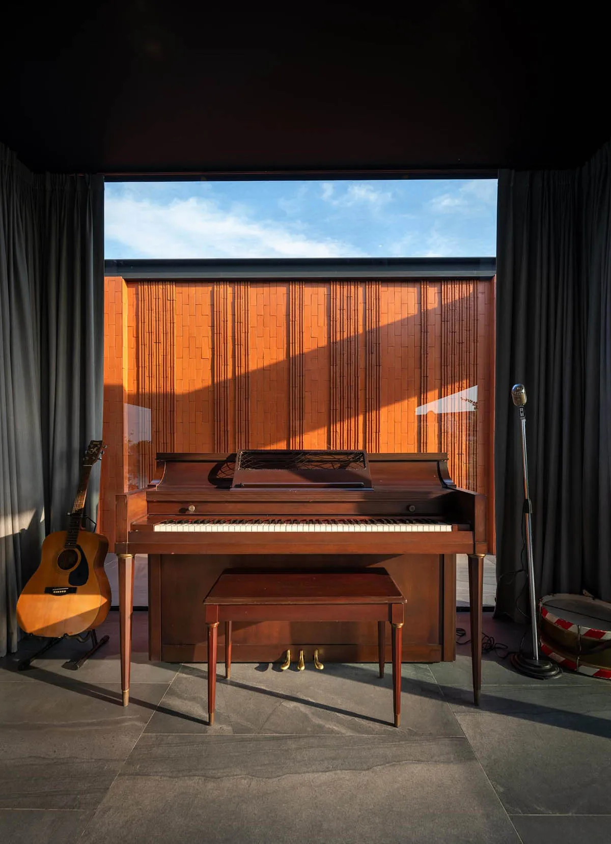 A minimalist music studio with wooden elements, featuring a piano, guitar, and microphone against a bamboo blind backdrop and