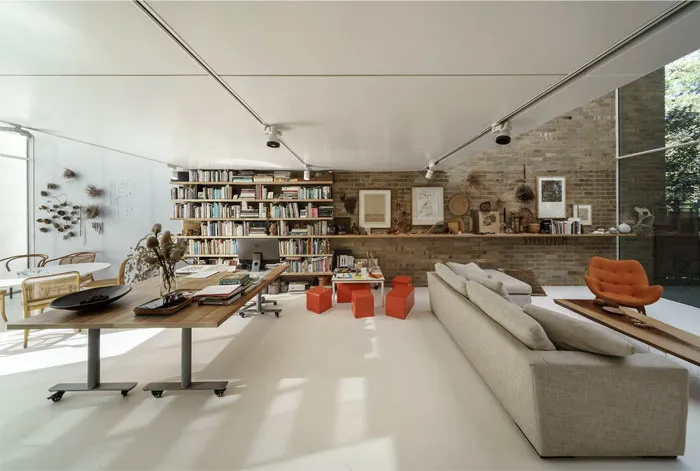 A minimalist living room with a concrete floor, a long sofa, red stools, and a bookshelf.