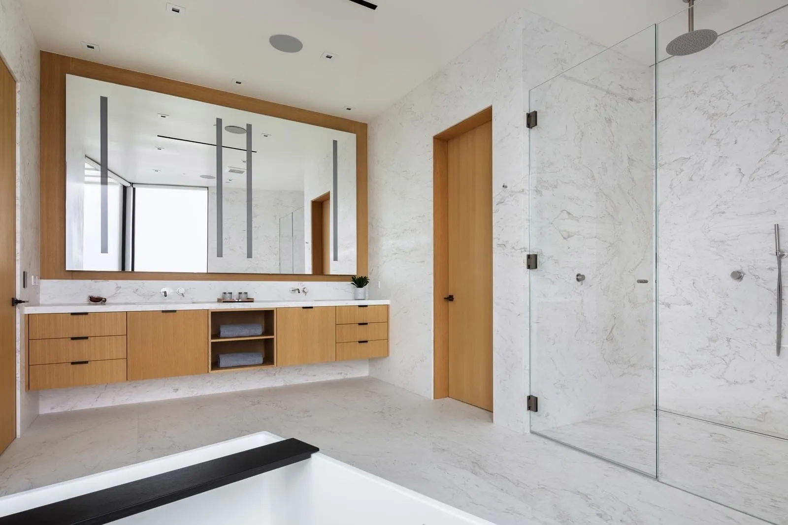 A modern bathroom with white marble walls, countertops, and floor tiles, featuring a large mirror, wooden vanity, and a glass